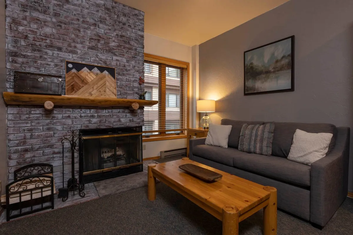 The image features a cozy interior with a wood-burning brick fireplace as the focal point, a plush grey sofa adorned with pillows, a wooden coffee table, and a softly glowing table lamp, creating a warm and inviting atmosphere perfect for relaxation after a day in the Colorado mountains.