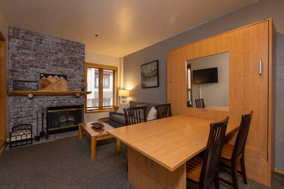 A warm and welcoming living room scene featuring a wood-burning fireplace with a stone surround, a fold-out wooden dining table, a comfortable sofa adorned with pillows, and a large window with wooden blinds that lets in natural light. The furniture arrangement creates an inviting space for relaxation, conversation, and shared meals, highlighting the heart of the home.