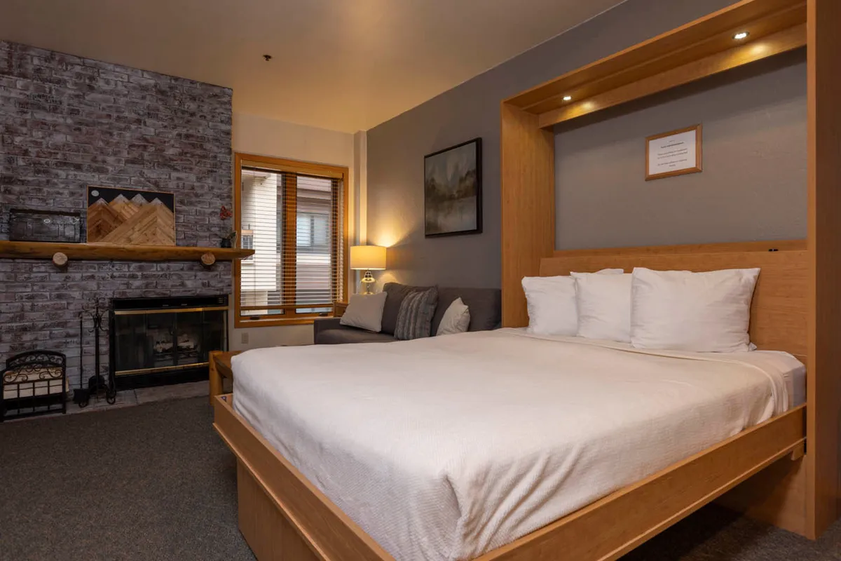 A cozy bedroom with a large, comfortable bed dressed in soft white linens sits near a rustic brick wood-burning fireplace. A small sitting area with a sofa and throw pillows offers space for relaxation, while a window with wooden blinds lets in gentle natural light, enhancing the tranquil ambiance.