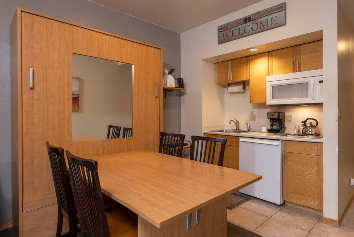 A cozy dining area with a wooden dining table and chairs, a compact kitchenette with sink and appliances, a large mirror embedded in a wooden wall unit, and a welcome sign above the kitchenette. The space encourages gathering for meals, conversation, or relaxing with a cup of coffee.