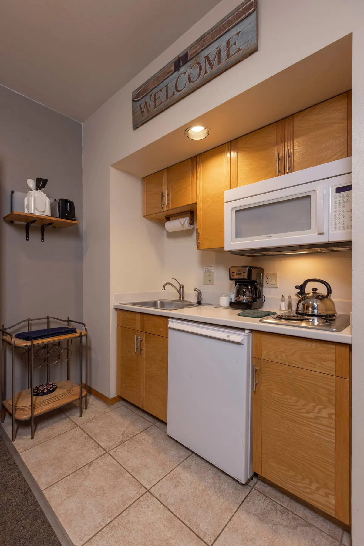 A beautifully lit kitchen with wooden cabinetry, a compact white microwave above the stove, a coffee maker and kettle on the countertop, and a decorative 'WELCOME' sign above the kitchen area. The setup is practical and homey, encouraging guests to prepare meals and feel immediately at ease upon arrival.