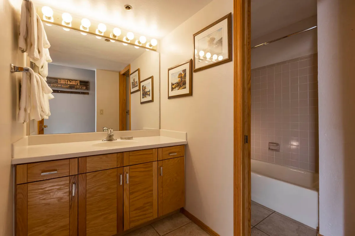 A well-lit bathroom featuring a large mirror with vanity lights, wooden cabinetry with a sink, and a tiled bathtub area visible through a sliding door. Decorative framed artwork adds a touch of personality and warmth to the practical, clean space.