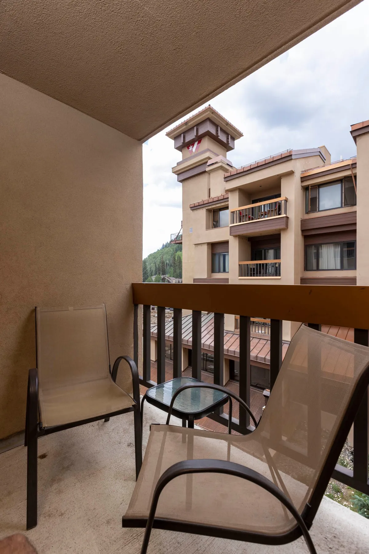 This image shows two beige mesh chairs and a small glass-top table set on a private balcony, with a view of an adjacent lodge building and its balconies. The setting is calm and intimate, suggesting a private space for couples to relax, enjoy mountain air, and share quiet moments together.