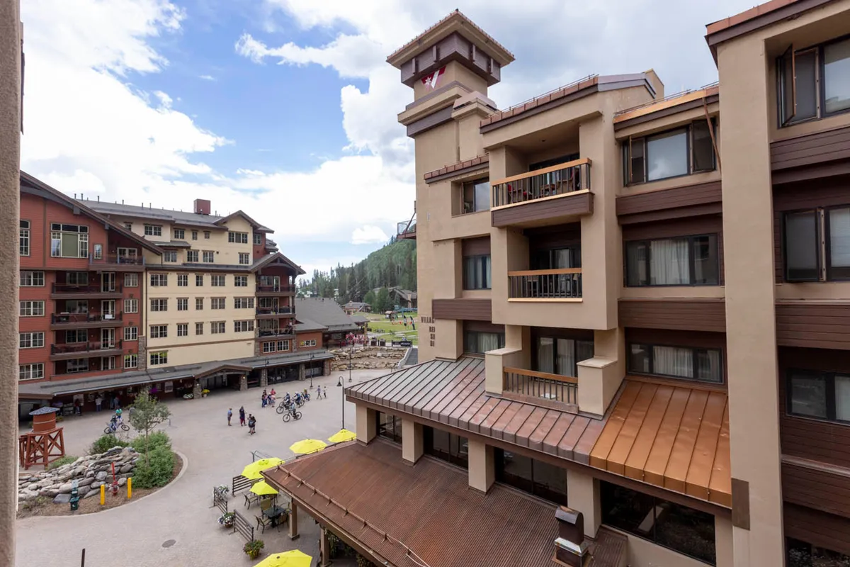 A vibrant view of the Village Plaza with people walking and biking, surrounded by lodge buildings. Yellow umbrellas and outdoor seating areas create a lively, social atmosphere, while the distant mountains set a scenic backdrop.