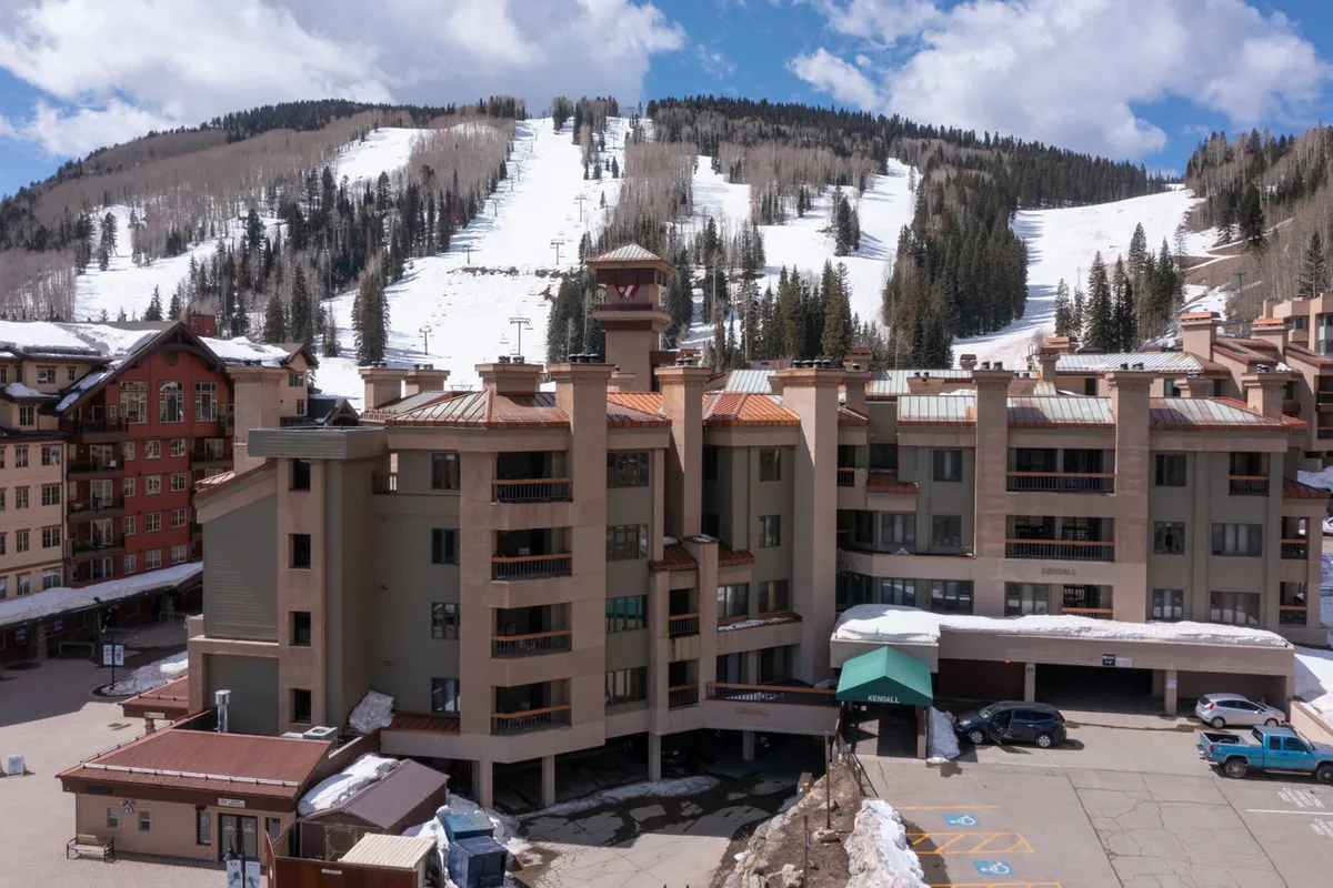 The Kendall Mountain Lodge building with snow-covered ski slopes rising behind and a parking area in front. The setting shows easy access from vehicle to lodge, with the surrounding mountain scenery adding to the resort vibe.