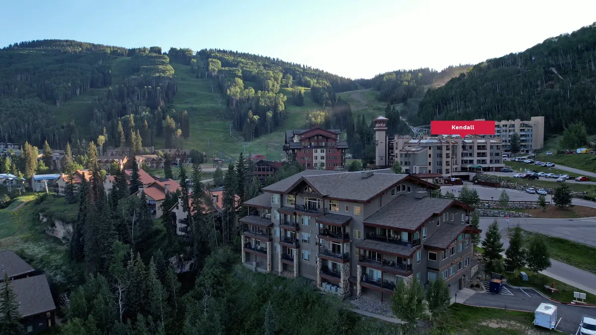 A wide exterior view captures the Kendall Mountain Lodge set against a backdrop of mountain slopes and forests, with nearby buildings and parking visible. The scene is tranquil and inviting, suggesting opportunities for outdoor adventure as well as relaxation.