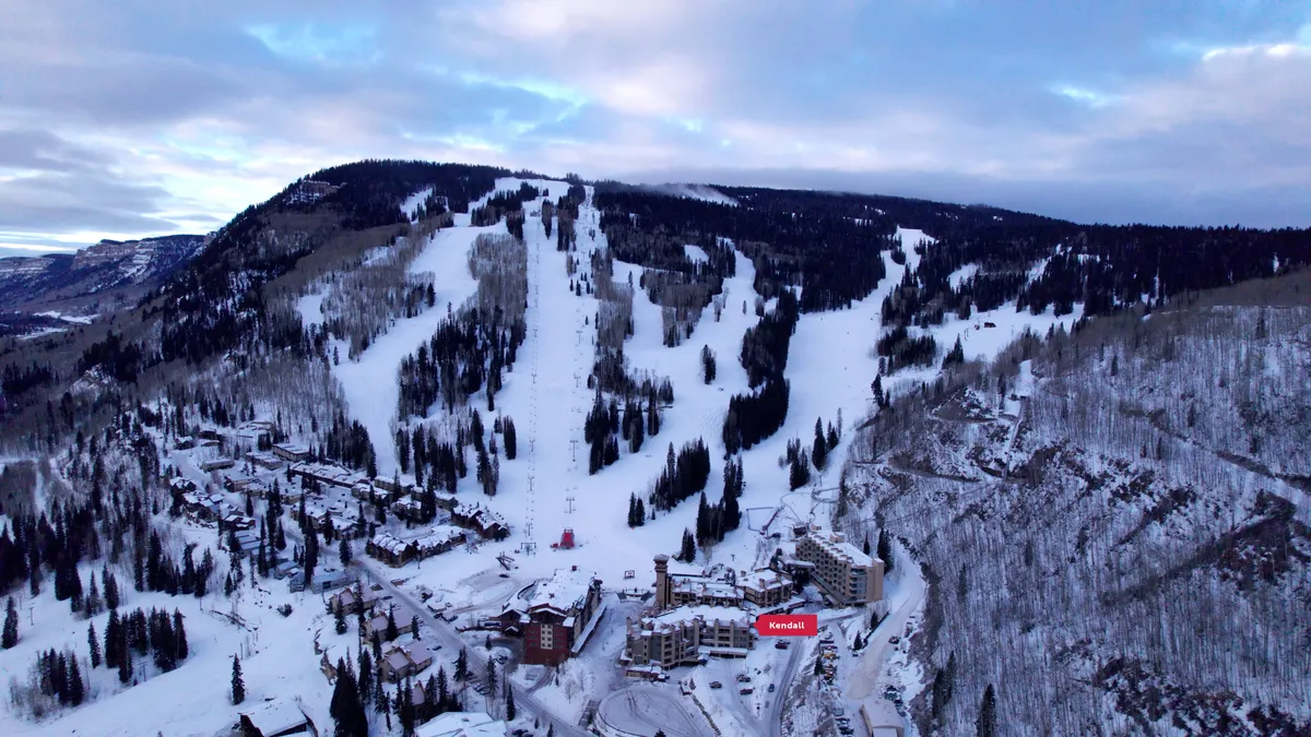 A panoramic winter view of the resort area, featuring snow-covered ski slopes and trails, ski lifts stretching up the mountain, and a cluster of resort buildings nestled among forested mountain terrain. Visitors can be seen skiing, snowboarding, and wandering through the nearby village plaza, with the majestic San Juan Mountains as a backdrop.