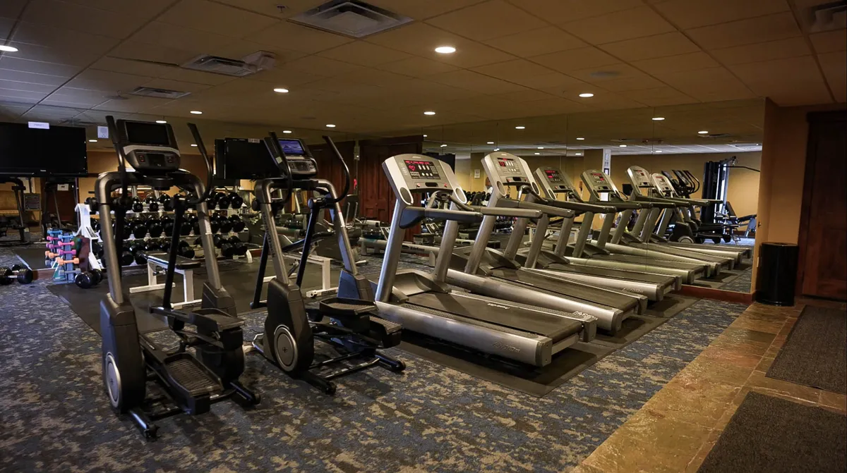A modern fitness center interior featuring a row of treadmills and elliptical machines, a free weights rack, and a mirrored wall. The space is well-lit, clean, and motivating, designed for guests seeking an energizing workout during their stay.