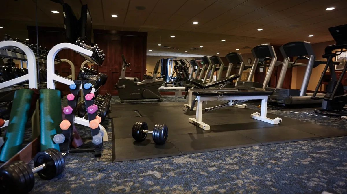 A bright, modern fitness room with dumbbells, a weight bench, treadmills, and exercise mats. The setup encourages both quick workouts and longer fitness routines.