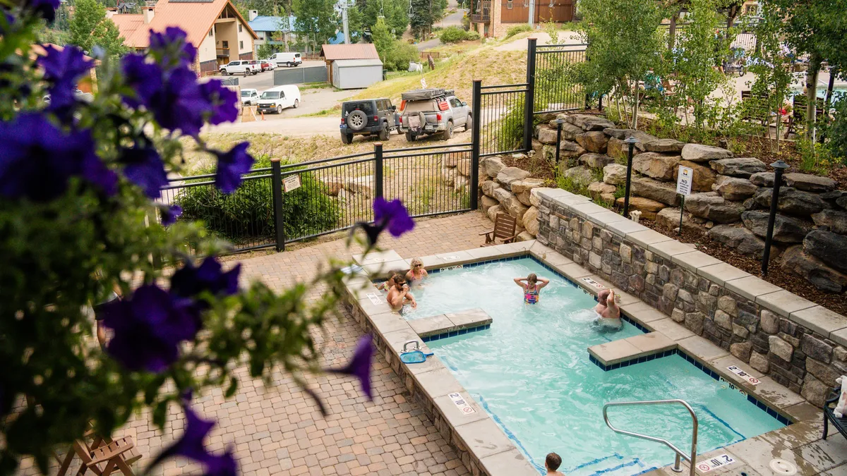 People are seen enjoying a heated outdoor swimming pool, enclosed by a stone wall and surrounded by a patio. Bright purple flowers in the foreground add a splash of color and vibrancy, while the metal safety gate ensures a secure, family-friendly environment. The atmosphere is lively and sociable, with groups relaxing and interacting poolside.