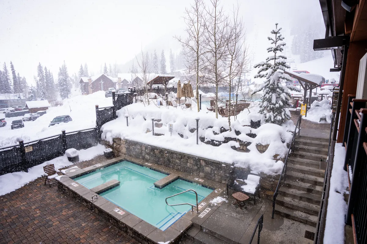 A heated outdoor swimming pool with vivid turquoise water is surrounded by a snow-draped stone wall and landscape. Steps lead up to an upper patio or hot tub area, while snow-blanketed pine trees and mountain chalets are visible in the background. The scene captures the striking contrast between the inviting warmth of the pool and the crisp, snowy environment.