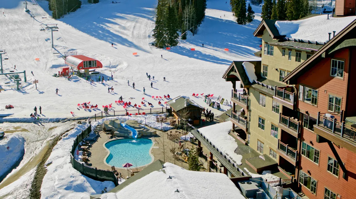 A steaming heated outdoor pool sits at the edge of a snow-dusted terrace, with guests swimming and relaxing poolside. In the background, skiers traverse the nearby slopes and a ski lift ascends the mountain, while the lodge building provides a cozy focal point.