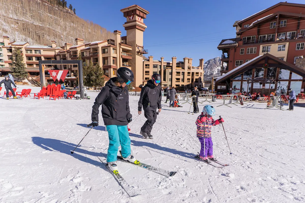 A vibrant outdoor scene featuring a family skiing together on fresh snow, with large, multi-story lodge buildings in the background. Red Adirondack chairs are arranged near the lodge, offering seating with mountain views. The mountain landscape creates a dramatic natural backdrop, and people can be seen walking around the resort plaza, suggesting convenient access to amenities.